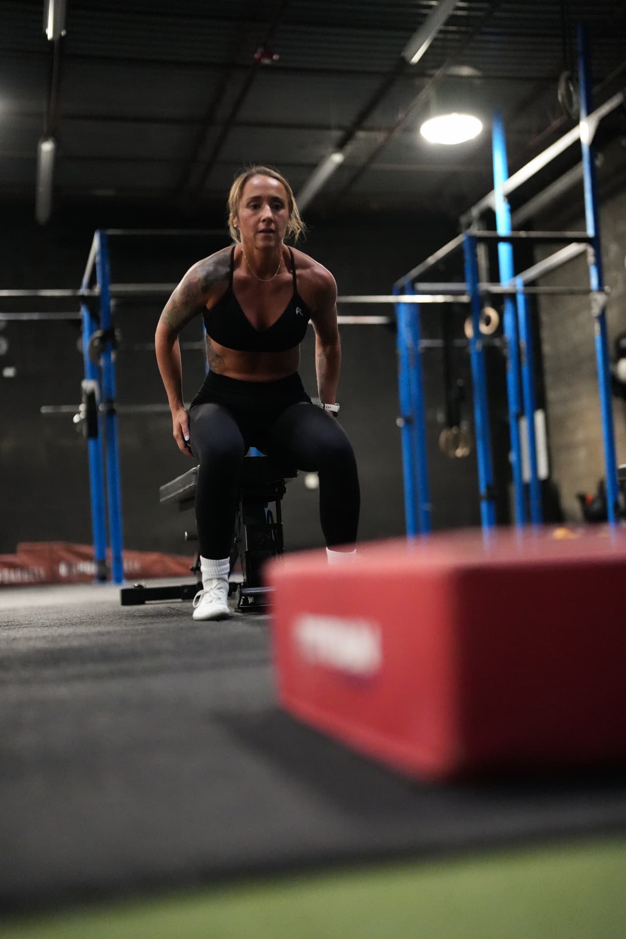 Woman seated on a weight bench in the gym, focused and ready for her next set with blue squat racks behind her.